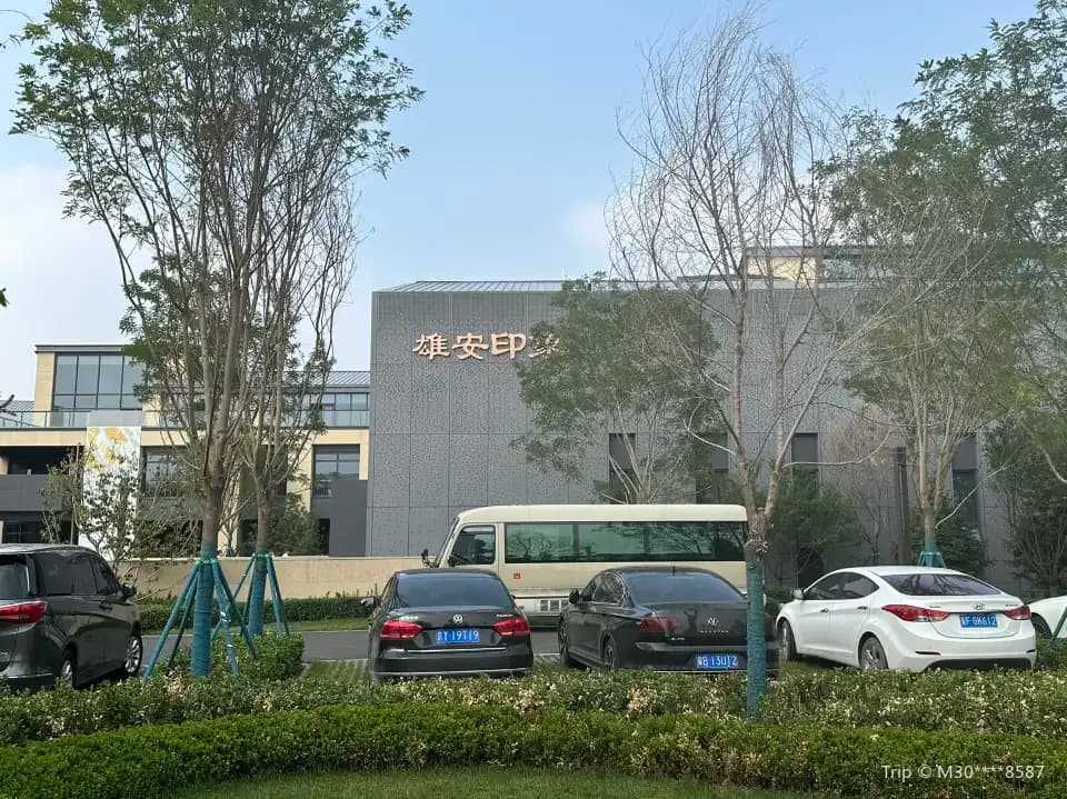 Das Ausstellungsgebäude von Xiong’an Impression hinter Bäumen; vor der Fassade stehen geparkte Fahrzeuge.